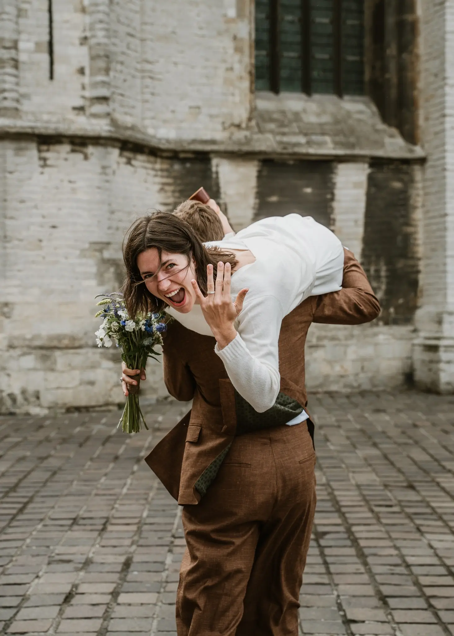 Wedding photography by Bert Van Den Eeckhaut — groom carrying bride over cobblestone street